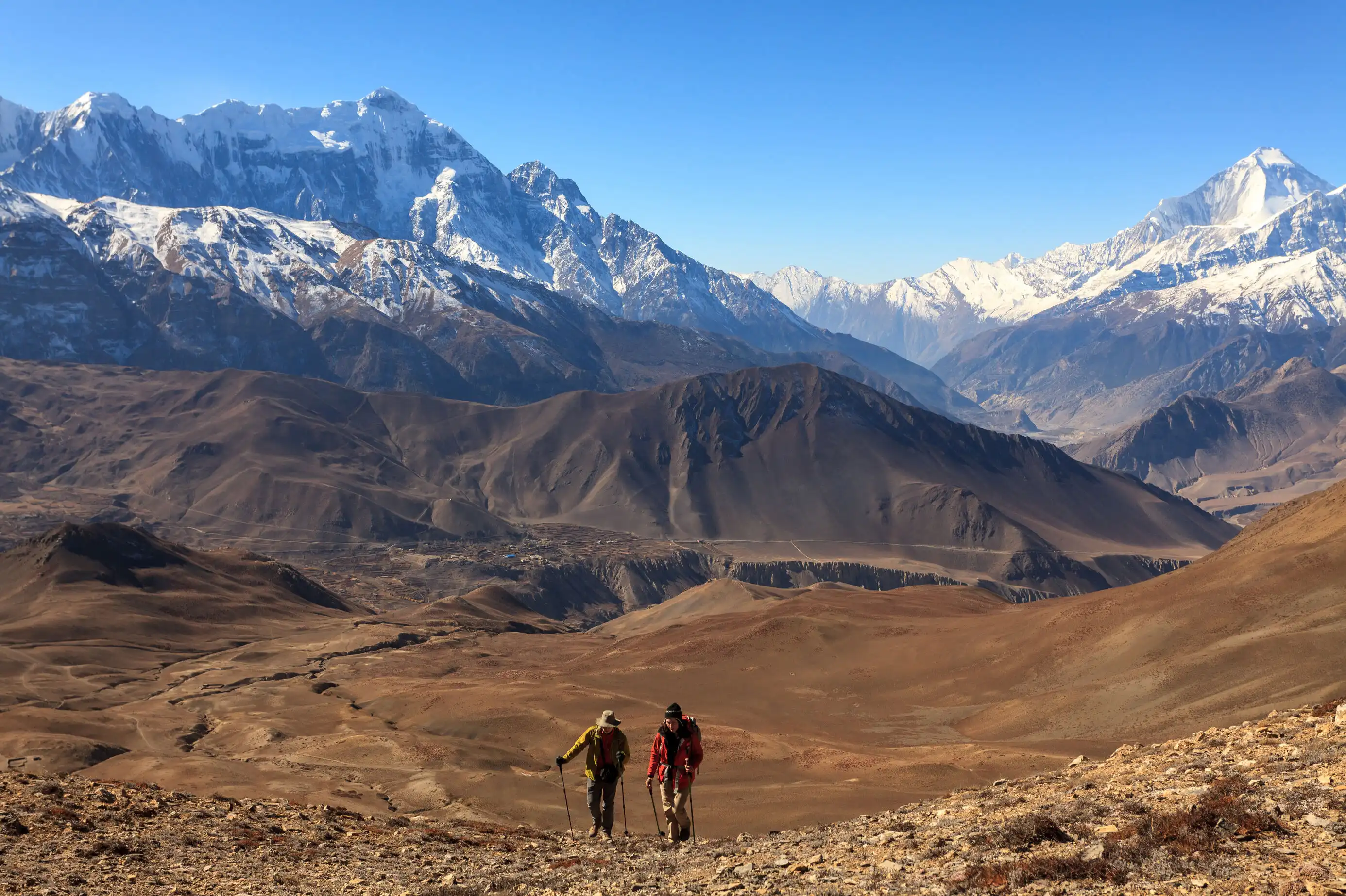 Two hikers trekking a rocky landscape with distant snow-covered mountains under a clear sky.