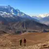 Two hikers trekking a rocky landscape with distant snow-covered mountains under a clear sky.
