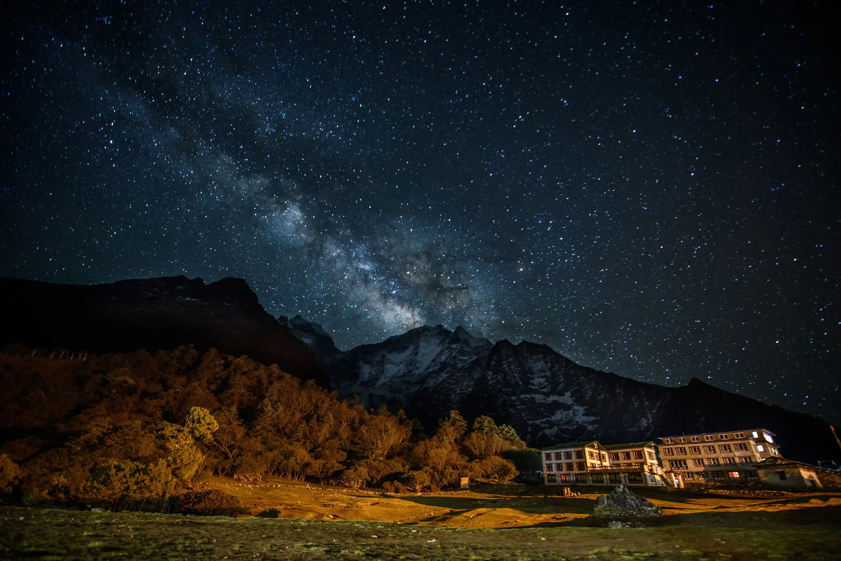 Milky Way stretching across a starry night sky above mountains, with a brightly lit building at the base.