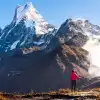 Hiker in bright pink jacket with trekking pole gazing at snowy Mardi Himal mountain under clear blue sky and mist.