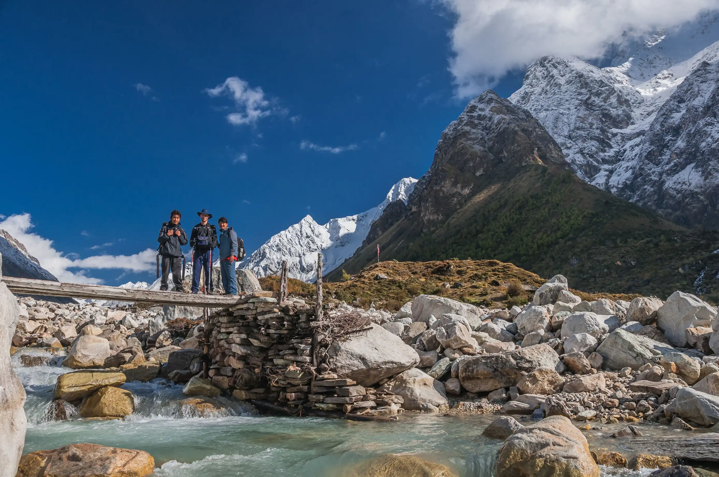Hikers crossing a wooden bridge over a clear stream with snow-capped mountains in the background.