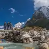 Hikers crossing a wooden bridge over a clear stream with snow-capped mountains in the background.