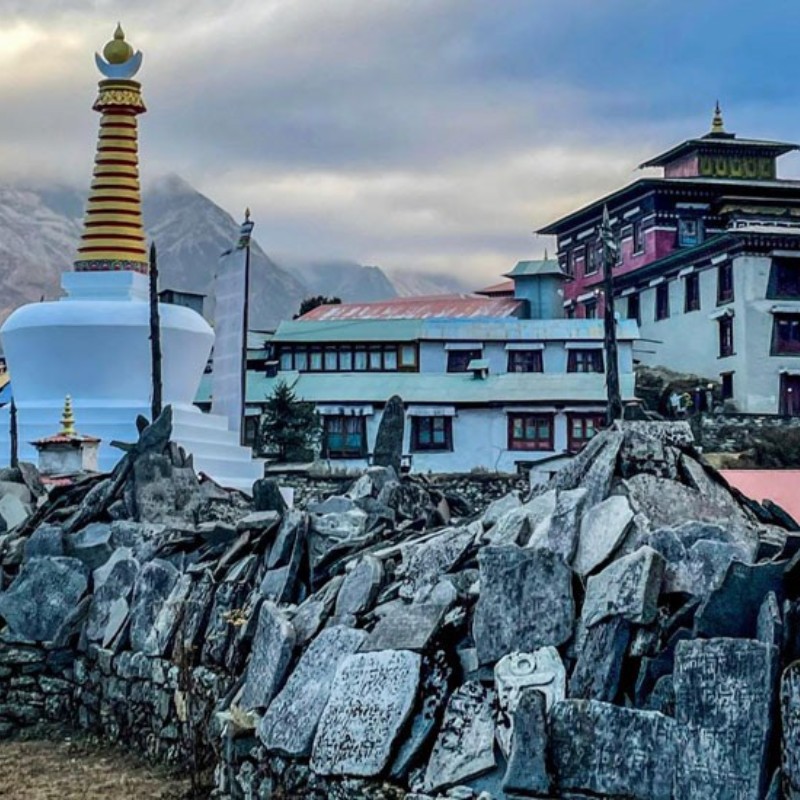 A Himalayan monastery complex with a large white stupa topped by a gold-and-red spire in the foreground, surrounded by stone walls and prayer-marked rocks. Behind it are multi-story monastery buildings painted in white, red, and blue, set against misty mountain peaks under a cloudy sky.
