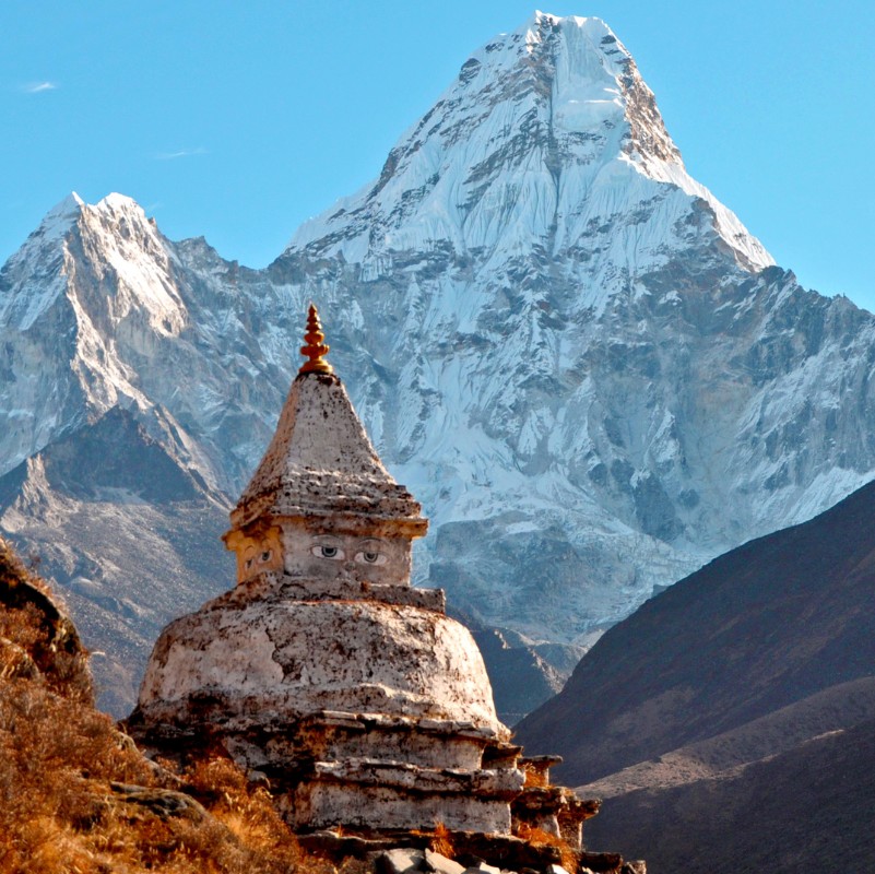 A traditional Buddhist stupa in the Everest region stands in the foreground with the towering snow-covered peak of Ama Dablam rising dramatically behind it under a clear blue Himalayan sky.