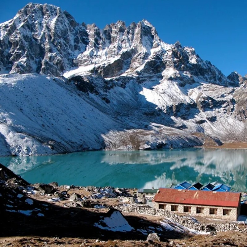 Turquoise Gokyo Lake surrounded by snow-covered Himalayan peaks under a clear blue sky, with mountain lodges and rocky terrain along the shoreline reflected in the still water.