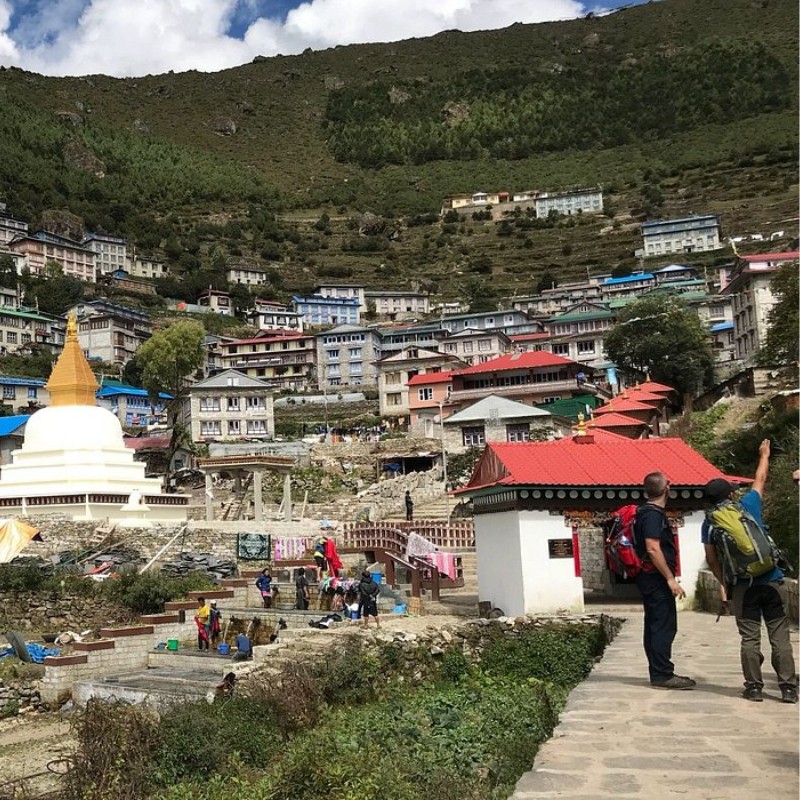 The entrance area of Namche Bazaar with a white Buddhist stupa, traditional buildings with colorful roofs, and trekkers standing on a stone pathway. The hillside town rises in terraces against a backdrop of green mountains and scattered clouds.