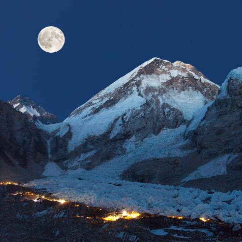 Nighttime view of the Khumbu Glacier beneath a snow-covered Himalayan peak, illuminated by moonlight and scattered camp lights along the glacier.