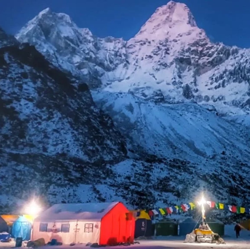 Snow-covered Ama Dablam rises dramatically under the blue twilight sky, its jagged peaks illuminated by the last light of day. In the foreground, a brightly lit red-and-white mountain camp sits on the snowy landscape, with colorful prayer flags strung across the scene and glowing lamps adding warmth to the cold Himalayan night.