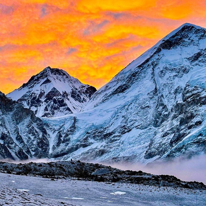 Sunrise over snow-covered Himalayan peaks near Kala Patthar, with glowing orange and pink clouds illuminating icy slopes, glaciers, and rugged mountain ridges in the foreground.