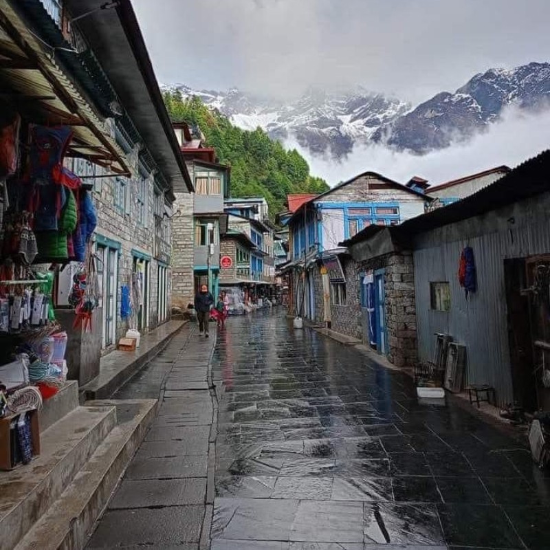 Wet street in Lukla market with shops and snow-covered mountains in background