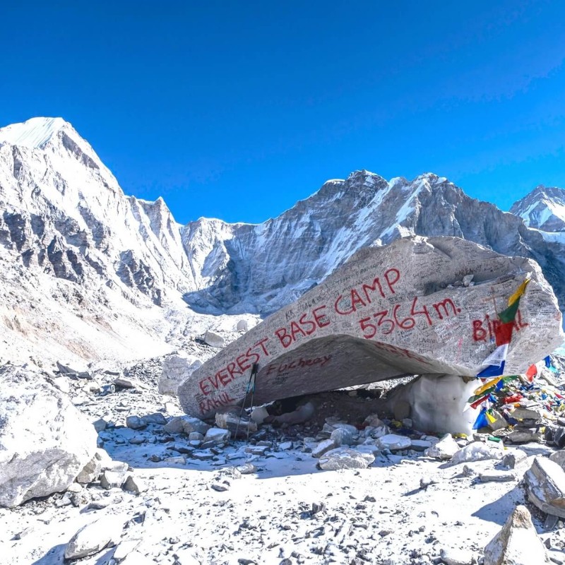 Snow-covered Himalayan landscape at Everest Base Camp with a large rock in the foreground painted with red graffiti reading “EVEREST BASE CAMP 5364m.” Prayer flags hang on the right side, and towering, rugged, snow-dusted peaks rise under a clear blue sky.