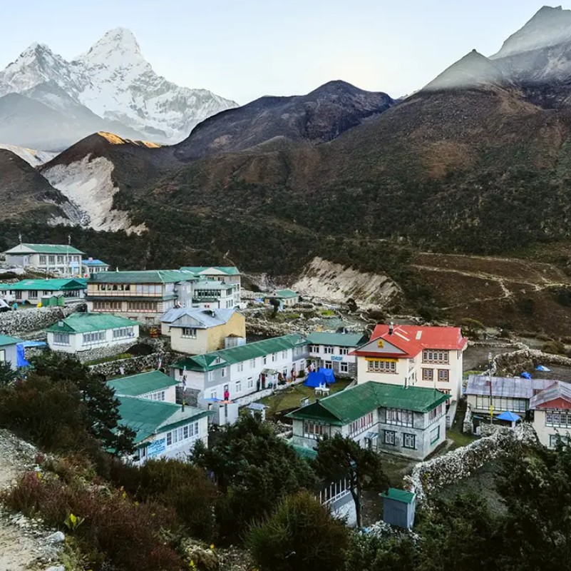 Pangboche village in the Everest region of Nepal, with lodges and teahouses nestled in a rugged mountain valley beneath the snow-covered peak of Ama Dablam, partially surrounded by clouds.